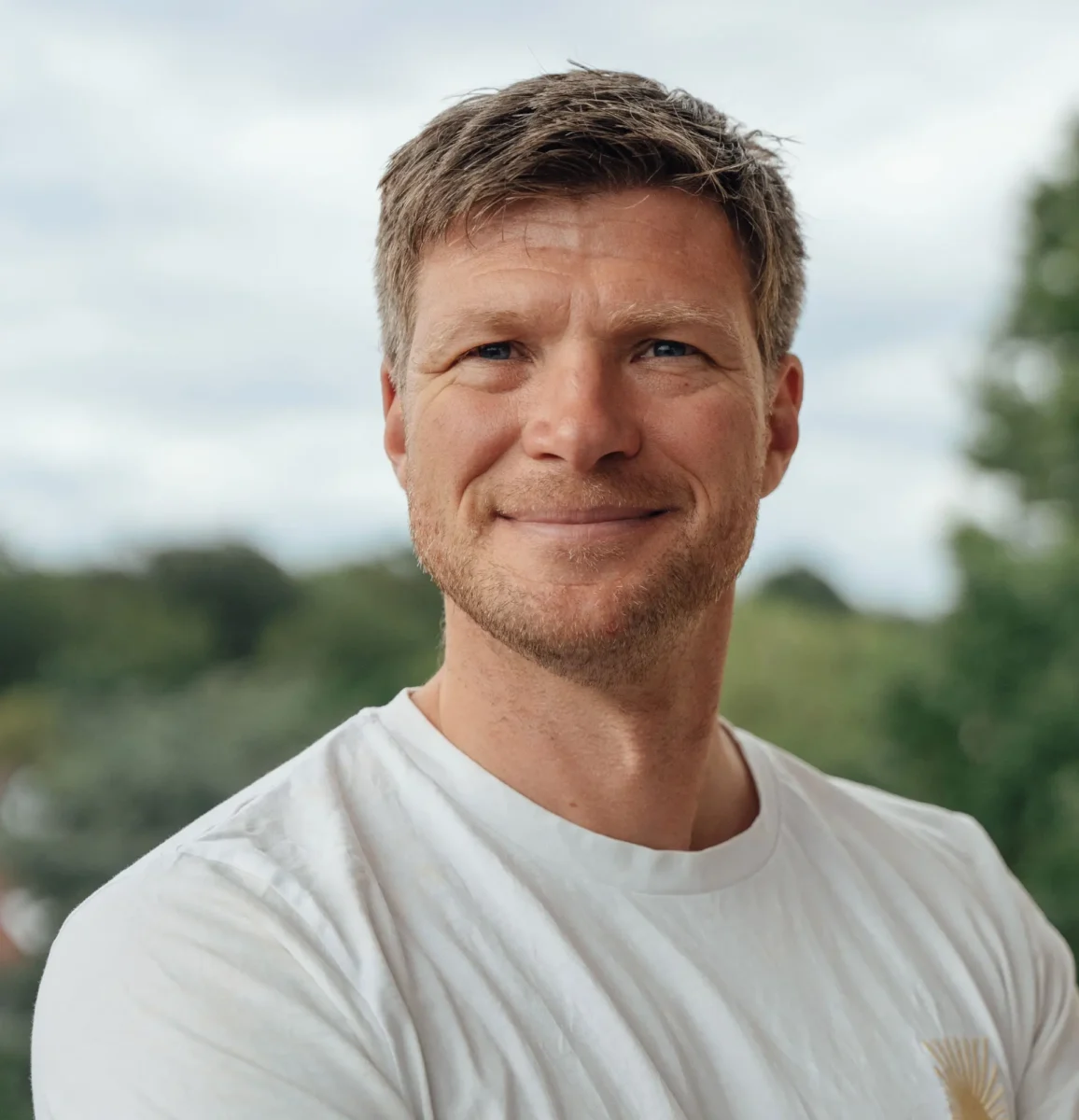 Ein Mann mit kurzen braunen Haaren und einem leichten Bart lächelt in die Kamera und trägt ein weißes T-Shirt. Der Hintergrund ist unscharf und zeigt Grünflächen und einen bewölkten Himmel. Er verkörpert die positiven Ergebnisse von „Mental Health Coaching Bremen“ und strahlt in seinem Alltag Wohlbefinden und Selbstvertrauen aus.