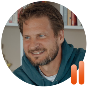 Ein Mann mit kurzen braunen Haaren und Bart, der einen blaugrünen Kapuzenpulli trägt, sitzt drinnen und lächelt. Im Hintergrund sind verschwommene Bücherregale zu sehen, die eine entspannte Sonntagsstimmung vermitteln. In der rechten unteren Ecke ist ein orangefarbenes Logo mit zwei Balken zu erkennen, das auf „Performance Coaching Bremen“ oder „Betriebliche Gesundheitsförderung Bremen“ hindeutet.