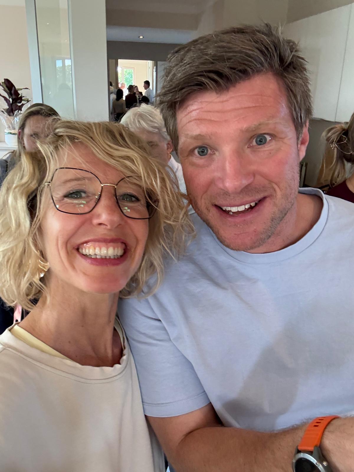 Zwei Personen lächeln und machen drinnen ein Selfie. Die Frau hat kurzes blondes Haar und trägt eine Brille; der Mann hat kurzes braunes Haar und trägt ein helles Hemd. Im Hintergrund sind weitere Personen und hell beleuchtete Räume zu sehen.