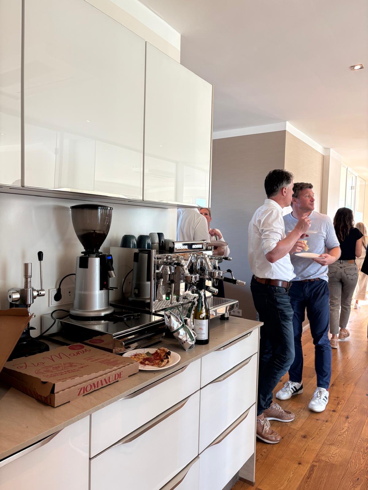 In einer hellen Küche stehen Menschen in der Nähe einer Kaffeemaschine und Pizzakartons und unterhalten sich. Auf der Arbeitsplatte stehen Teller mit Pizzastücken und Getränke. Der Boden ist aus Holz, und Sonnenlicht durchflutet den Raum.