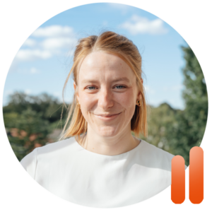 Eine Frau mit hellbraunen Haaren und einem weißen Oberteil lächelt an einem sonnigen Sonntag im Freien, umrahmt von Bäumen und einem blauen Himmel. In der rechten unteren Ecke des Bildes sind zwei vertikale orangefarbene Balken zu sehen, die auf subtile Weise die lebendige Energie von Performance Coaching Bremen vermitteln.