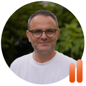Ein Mann mit kurzen dunklen Haaren, Brille und weißem Hemd lächelt im Freien vor grünem Laub – und fängt so die frische Stimmung eines Sonntags ein. Unten rechts im runden Rahmen ist eine orangefarbene Grafik mit zwei Balken zu sehen, die für Performance Coaching Bremen steht.